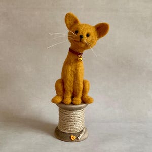 May include: A handmade, orange felt cat figure with a red collar and a small gold bell. The cat sits on a wooden spool wrapped with twine. The cat has long white whiskers. A whimsical decorative item.