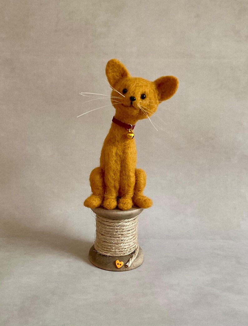 May include: A handmade, orange felt cat figure with a red collar and a small gold bell. The cat sits on a wooden spool wrapped with twine. The cat has long white whiskers. A whimsical decorative item.