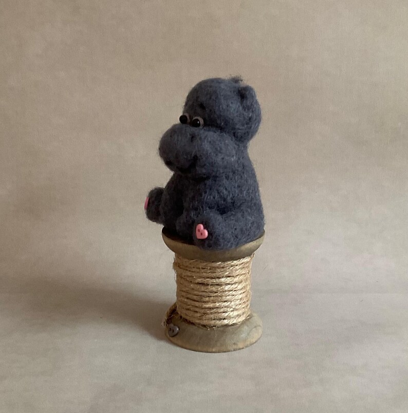 May include: A dark grey felt hippo figure with pink details, sitting on a wooden spool wrapped with twine. The hippo has small black eyes and pink feet. The background is a neutral tan colour.