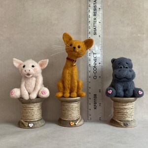 May include: Three handmade felt animal figurines: a pink pig, an orange cat with a red collar, and a grey hippo. Each animal sits atop a wooden spool. The ruler next to the animals measures up to 53 cm.