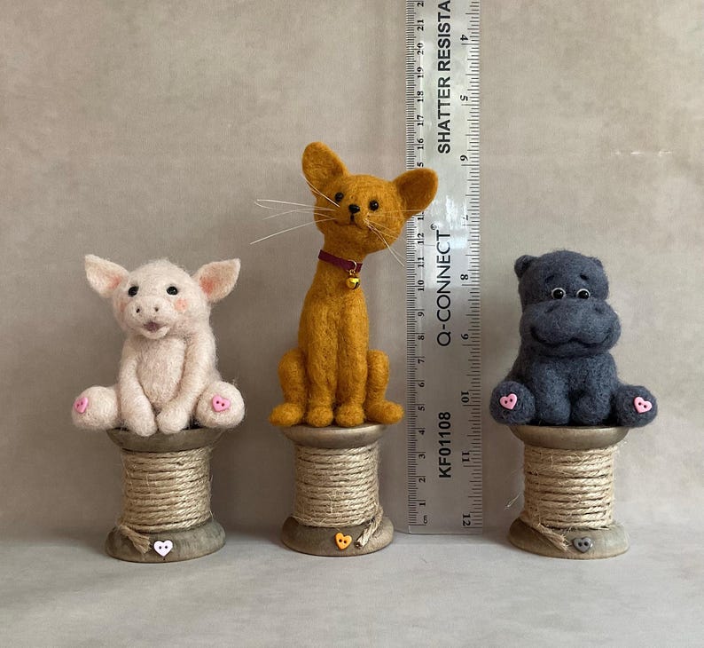 May include: Three handmade felt animal figurines: a pink pig, an orange cat with a red collar, and a grey hippo. Each animal sits atop a wooden spool. The ruler next to the animals measures up to 53 cm.