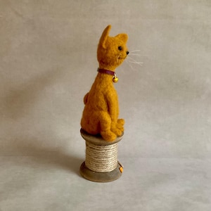 May include: An orange cat figurine with a purple collar and a gold bell, sitting on a wooden spool wrapped with twine. The cat has visible whiskers. A handmade decorative item. The spool is a charming base for the cat.