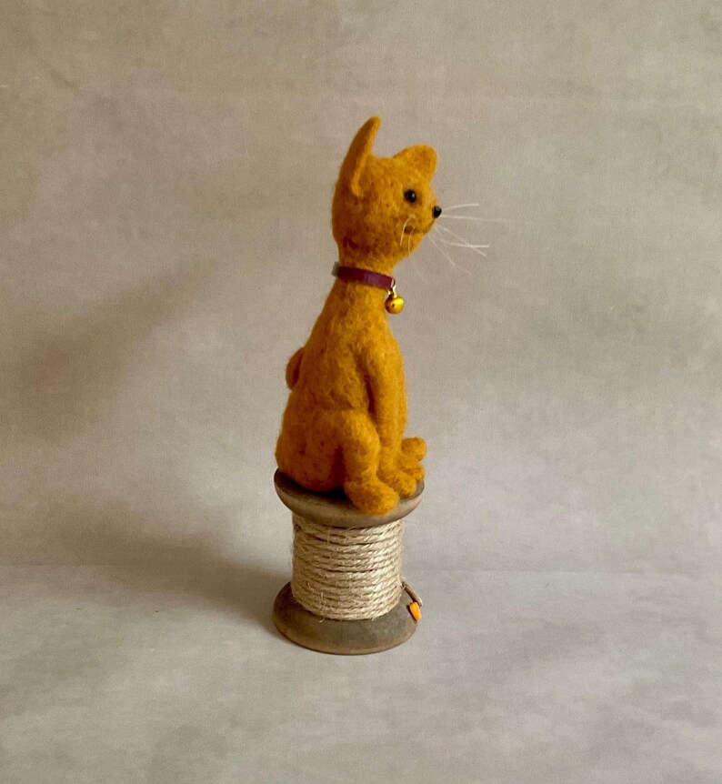 May include: An orange cat figurine with a purple collar and a gold bell, sitting on a wooden spool wrapped with twine. The cat has visible whiskers. A handmade decorative item. The spool is a charming base for the cat.