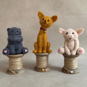 May include: Three handmade felt animal figurines: a dark blue hippo, a yellow cat with a bell collar, and a pink pig. Each animal sits atop a wooden spool with a heart-shaped button.