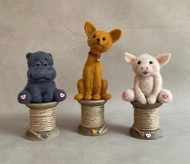 May include: Three handmade felt animal figurines: a dark blue hippo, a yellow cat with a bell collar, and a pink pig. Each animal sits atop a wooden spool with a heart-shaped button.