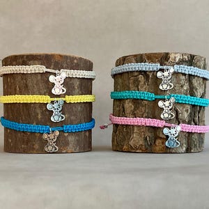 May include: Macramé bracelets in beige, yellow, and blue, and in light blue, teal, and pink. Each bracelet features a silver-coloured mouse charm. The bracelets are displayed on sections of tree trunks against a neutral background.
