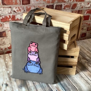 May include: A grey canvas tote bag with a stack of three embroidered frogs. The frogs are pink, purple and blue.