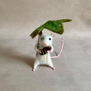 May include: A handcrafted felt mouse figurine, holding a brown and black object, with a green leaf overhead. The mouse has a white body, pink ears, and a pink tail. The leaf has water droplets.