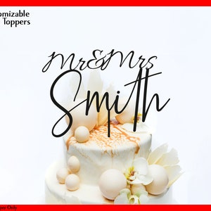 May include: White wedding cake with a black acrylic cake topper that says "Mr & Mrs Smith" in a cursive font.