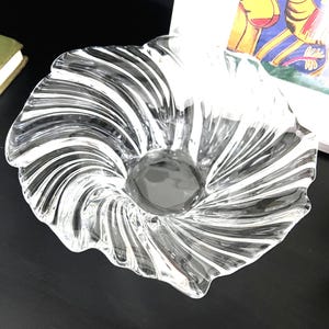 May include: A clear glass bowl with a swirling, ridged design. The bowl has a central depression and a scalloped edge. The glass is transparent, allowing light to pass through, and is set against a dark surface.