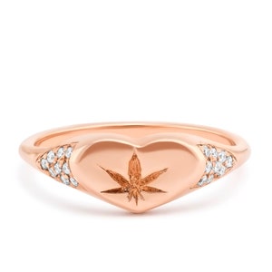 Puede incluir: Anillo de oro rosa con un engaste en forma de corazón engastado con un solo diamante y un diseño de hoja de cannabis. La banda está acentuada con diamantes pavé.