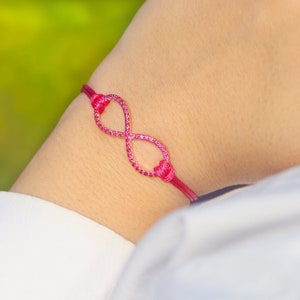 May include: A pink infinity symbol bracelet with small pink stones. The bracelet is tied on a person's wrist.