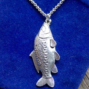 Op de afbeelding: Een zilveren ketting met een gedetailleerde zilveren vis hanger. De vis heeft een getextureerd oppervlak en hangt tegen een blauwe achtergrond.
