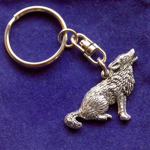 May include: Silver key chain with a wolf charm. The wolf is sitting with its head turned to the side and its mouth slightly open. The key chain is on a blue background.