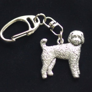 May include: Silver-colored keychain featuring a detailed poodle dog charm. The charm is attached to a metal clasp and a small chain. The dog charm has a textured surface and is standing on all fours.