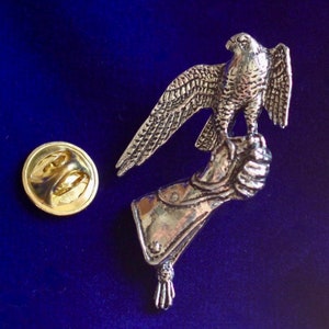 Może przedstawiać: Srebrna metalowa spinka z przedstawieniem sokoła siedzącego na ręce w rękawiczce. Spinka ma złote plecy z zapięciem bezpieczeństwa.