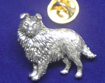 Puede incluir: Un broche de metal plateado con forma de perro collie de pelo largo. El broche presenta una textura de pelaje detallada y una pose de pie. Un cierre circular de metal dorado se encuentra posicionado directamente sobre la figura.