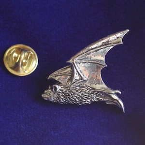 Pode incluir: Um broche prateado em forma de morcego, com asas abertas e detalhes texturizados. Ao lado do morcego, um fecho dourado. O broche está sobre um fundo azul escuro, ideal para temas góticos.