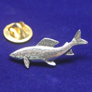 May include: A silver-toned metal lapel pin shaped like a fish, featuring detailed scales, fins, and a tail. The pin includes a separate gold-toned circular clutch back fastener, which is positioned slightly above and to the left of the fish figure.