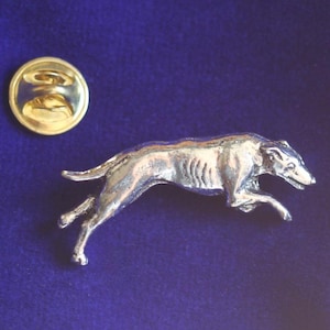 May include: A silver-toned metal lapel pin shaped like a greyhound dog in a running pose, featuring a detailed muscular body and a slender tail. The pin includes a separate circular gold-toned metal clutch back fastener with a central post.
