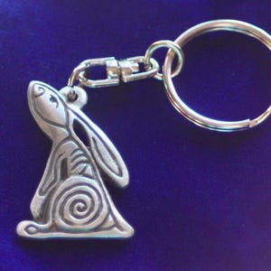 Puede incluir: Un llavero de color plateado con un amuleto de conejo detallado. El conejo está de pie sobre sus patas traseras con las orejas levantadas y tiene un diseño en espiral en la espalda.