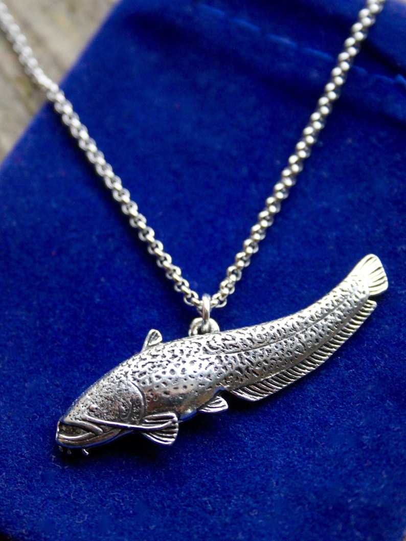 May include: A silver necklace with a detailed catfish pendant. The necklace is on a blue velvet background.