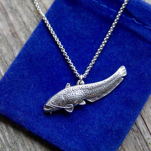 May include: A silver necklace with a detailed catfish pendant. The necklace is displayed on a blue velvet pouch.
