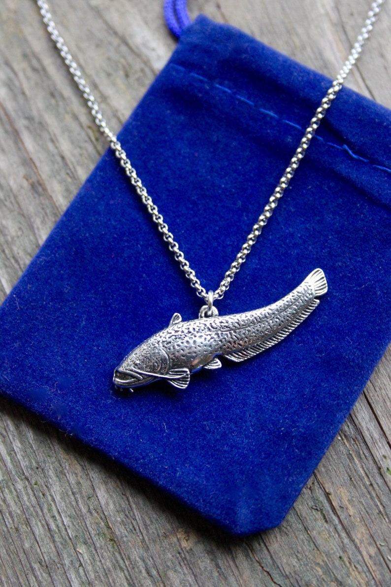 May include: A silver necklace with a detailed catfish pendant. The necklace is displayed on a blue velvet pouch.