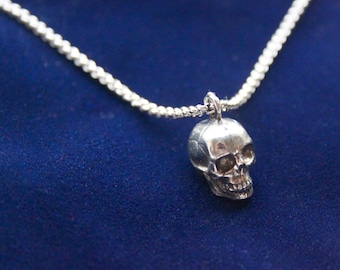 May include: A silver skull pendant necklace. The pendant is a detailed miniature skull with visible eye sockets and teeth. The necklace chain is a delicate, textured silver. The background is a deep blue.