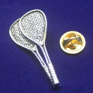Könnte beinhalten: Eine silberfarbene Anstecknadel aus Metall, die zwei gekreuzte Squashschläger mit detailliertem Saitenmuster und strukturierten Griffen zeigt, begleitet von einem separaten runden, goldfarbenen Metallverschluss zur Befestigung am Stoff.