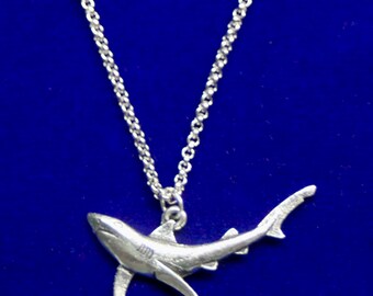 May include: A silver-toned shark pendant suspended from a delicate silver-toned metal chain. The shark charm is depicted in a swimming position with detailed fins and a textured body, hanging from a small jump ring attached to the chain links.