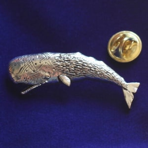 Puede incluir: Un broche de ballena plateada con detalles texturizados y boca abierta. El broche está junto a un broche redondo dorado con cierre sobre un fondo azul. El broche de ballena es un accesorio único.