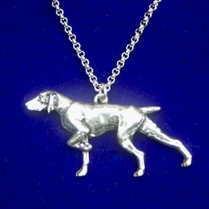 Può includere: Un ciondolo in metallo color argento a forma di cane da ferma, sospeso a una catena a maglie color argento abbinata. La figura del cane è raffigurata in una classica posa da caccia con una zampa anteriore sollevata, mostrando muscoli dettagliati.
