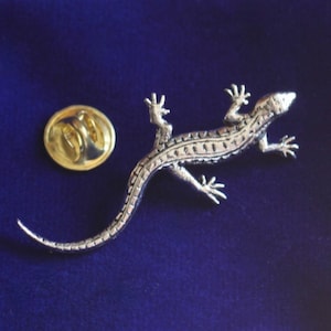Puede incluir: Pin de lagarto plateado con un diseño detallado. El pin tiene una parte posterior dorada con un cierre de seguridad.