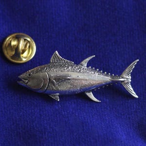 May include: A silver-colored tuna fish pin with detailed scales and fins, next to a gold-colored pin backing. The pin is set against a deep blue background, creating a striking contrast.