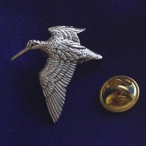 Könnte beinhalten: Eine silberfarbene Anstecknadel mit einem im Flug befindlichen Schnepfenvogel. Die Nadel hat eine goldfarbene Rückseite mit einem Sicherheitsverschluss.