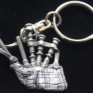 Può includere: Un portachiavi a forma di cornamusa argentata. La cornamusa è dettagliata con un motivo a quadri e un nastro.