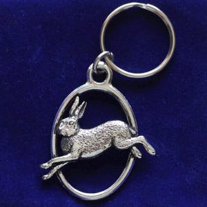 Puede incluir: Un llavero de plata con un anillo circular y un charm ovalado que presenta una liebre de plata saltando en un círculo.
