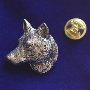 May include: A silver-toned metal lapel pin shaped like the head of a wolf in profile, featuring detailed fur texture and pointed ears. The pin includes a separate circular gold-toned metal clutch back fastener positioned to the right of the wolf head.