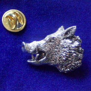 Könnte beinhalten: Eine silberfarbene Anstecknadel mit einem detaillierten, dreidimensionalen Eberkopf mit strukturierter Oberfläche. Der Eber hat den Mund offen und zeigt scharfe Zähne. Die Nadel hat eine goldfarbene Rückseite mit einer Sicherheitsnadel zum Verschließen.
