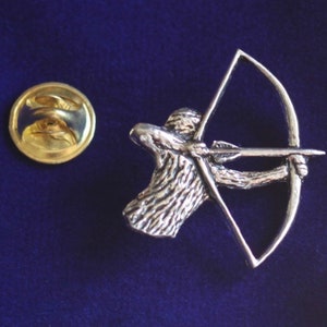 May include: Silver-toned pin featuring a detailed depiction of an archer with a bow and arrow. The pin has a gold-toned back with a safety pin closure.