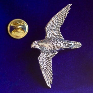 May include: A silver-coloured pewter pin of a falcon in flight. The pin has a gold-coloured back with a safety clasp.