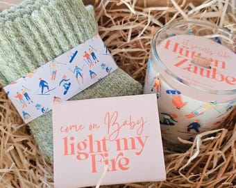 May include: A gift box containing a pair of light green socks with a skier design, a candle with the text "It smells like Hütten Zauber", and a small pink card with the text "come on Baby light my Fire". The box is filled with straw.