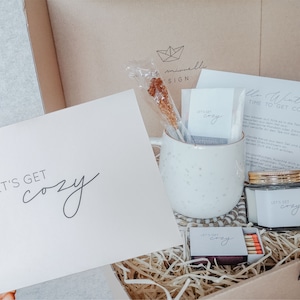 Könnte beinhalten: Eine Geschenkbox mit gemütlichem Thema, die eine weiß gesprenkelte Tasse, ein Glas mit goldenem Deckel, Streichhölzer und Karten enthält. Auf den Karten steht "Let's Get Cozy" und "Hello Winter, Time to Get Cozy". Ein Kandiszucker-Stiel steckt in der Tasse.