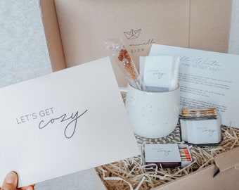 May include: A gift box with a cozy theme, containing a white speckled mug, a jar with a gold lid, matches, and cards. The cards read "Let's Get Cozy" and "Hello Winter, Time to Get Cozy". A rock candy stick is in the mug.