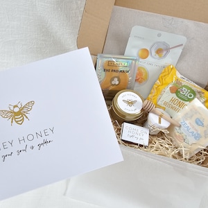 Könnte beinhalten: Eine Geschenkbox gefüllt mit Honig-Beauty- und Lebensmittelprodukten. Die Box enthält eine Karte mit dem Text "HEY HONEY your soul is golden", ein Honigglas, einen Honigstab, eine Honig-Augenmaske, eine Honigwaffel und eine Honig-Seife.