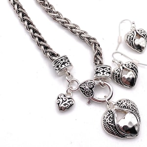 May include: A silver necklace with a braided chain and a heart pendant with wings. The pendant has a smaller heart inside. The necklace comes with matching earrings.