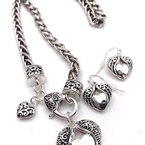 May include: A silver necklace with a heart pendant and matching earrings. The necklace has a braided chain and a heart-shaped clasp. The pendant is a large heart with a smaller heart inside. The earrings are small hearts with wings.