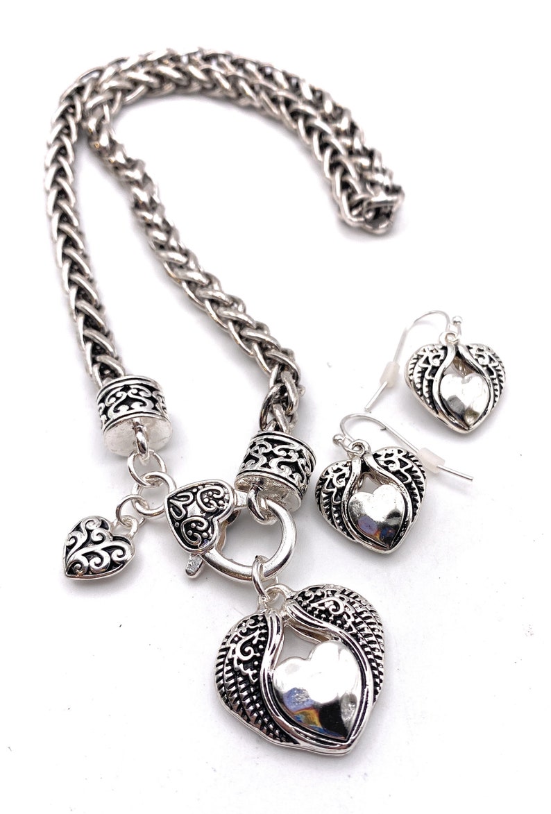 May include: A silver necklace with a heart pendant and matching earrings. The necklace has a braided chain and a heart-shaped clasp. The pendant is a large heart with a smaller heart inside. The earrings are small hearts with wings.