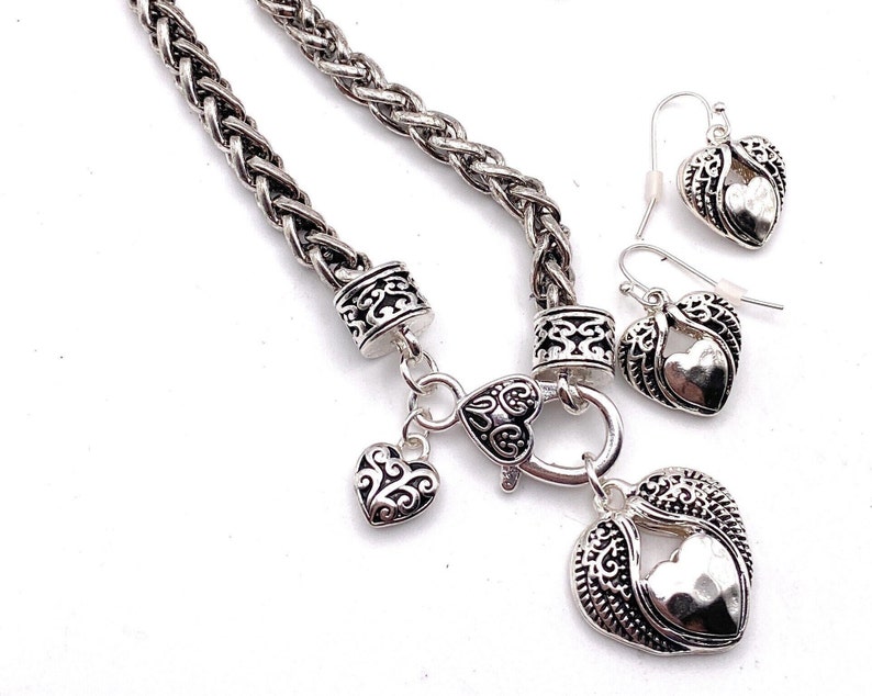 May include: A silver necklace with a braided chain and a heart pendant with wings. The pendant has a smaller heart inside. The necklace comes with matching earrings.